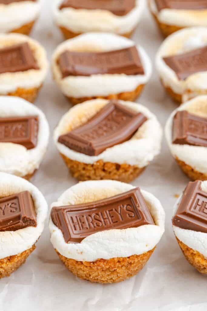 Rows of mini s'mores cups with graham crust, toasted marshmallow, and chocolate on parchment paper.