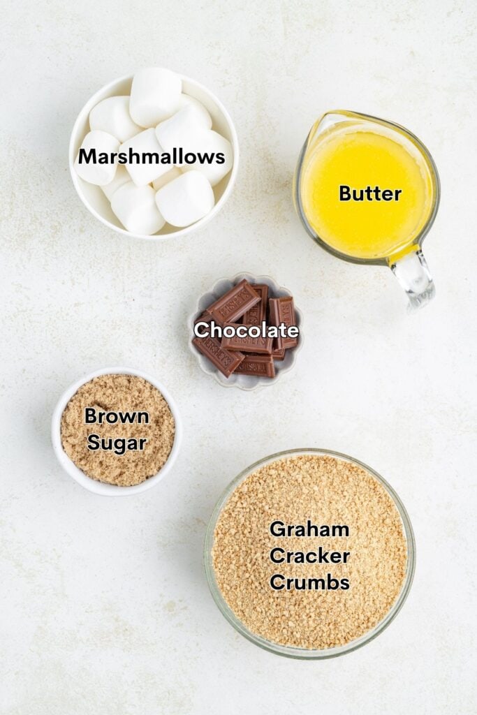 Five labeled ingredients—marshmallows, melted butter, chocolate, brown sugar, graham cracker crumbs—on a white surface.