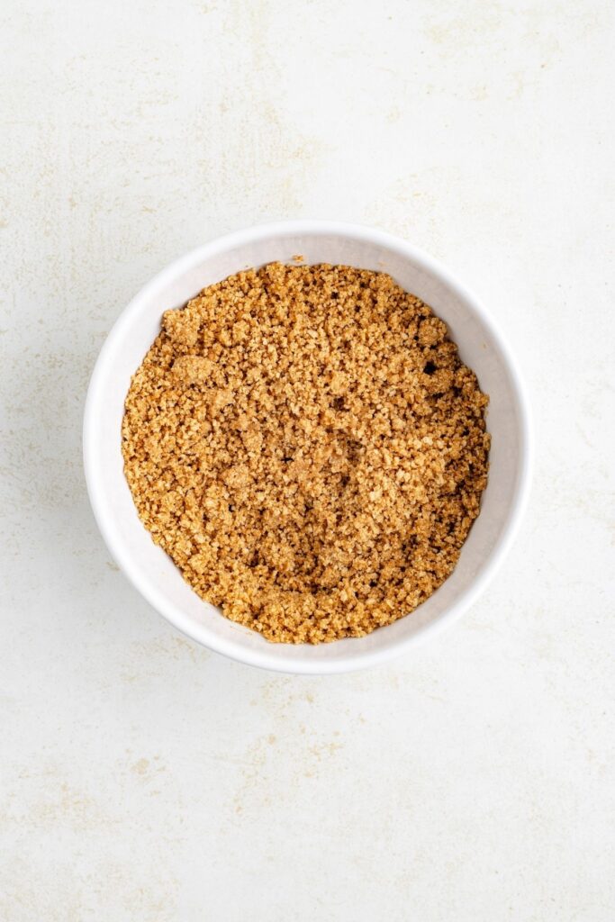 A white bowl of graham cracker crust mixture sits on a light, textured surface.