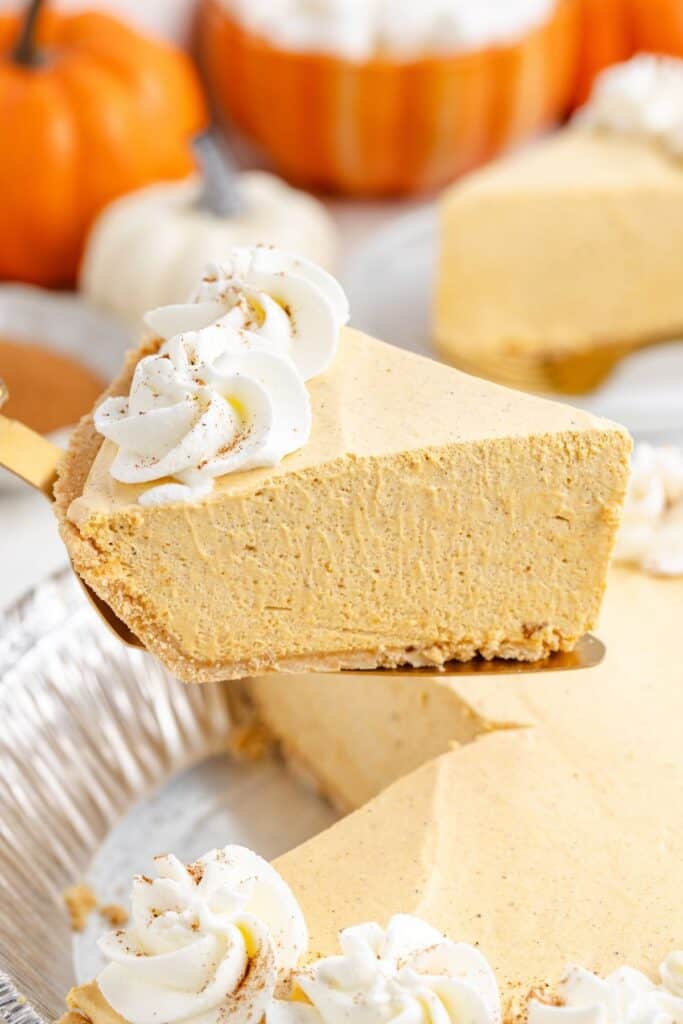 A slice of no-bake marshmallow pumpkin pie with graham cracker crust and whipped cream is lifted from a tin, pumpkins behind.