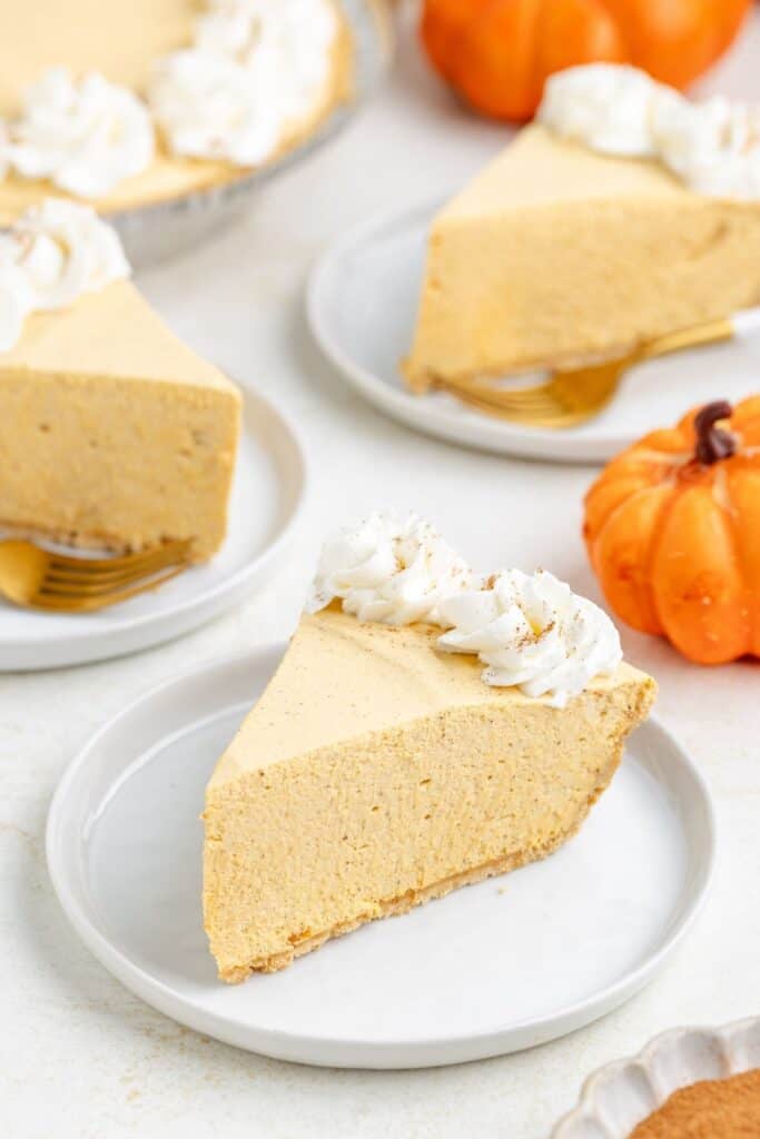 Slice of pumpkin marshmallow pie with whipped cream on a white plate, whole pie and small pumpkins behind.