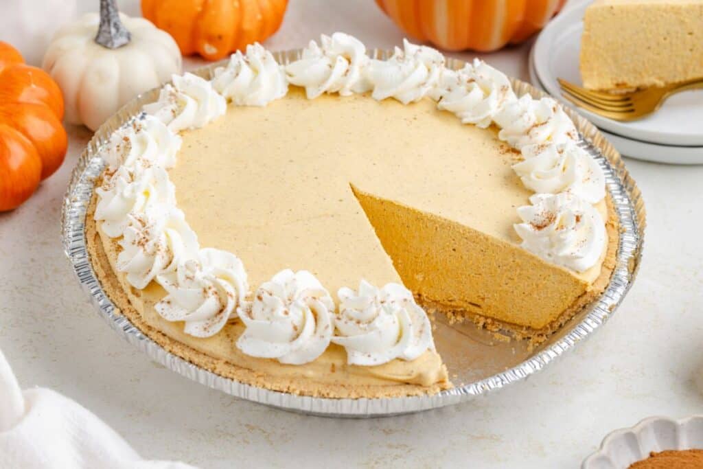 Marshmallow pumpkin pie with whipped cream on a table, one slice on a plate. Decorative pumpkins in background.
