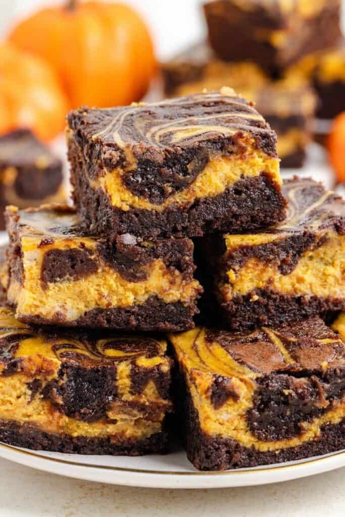Pumpkin swirl brownies stacked on a white plate, small orange pumpkins softly blurred in the background.