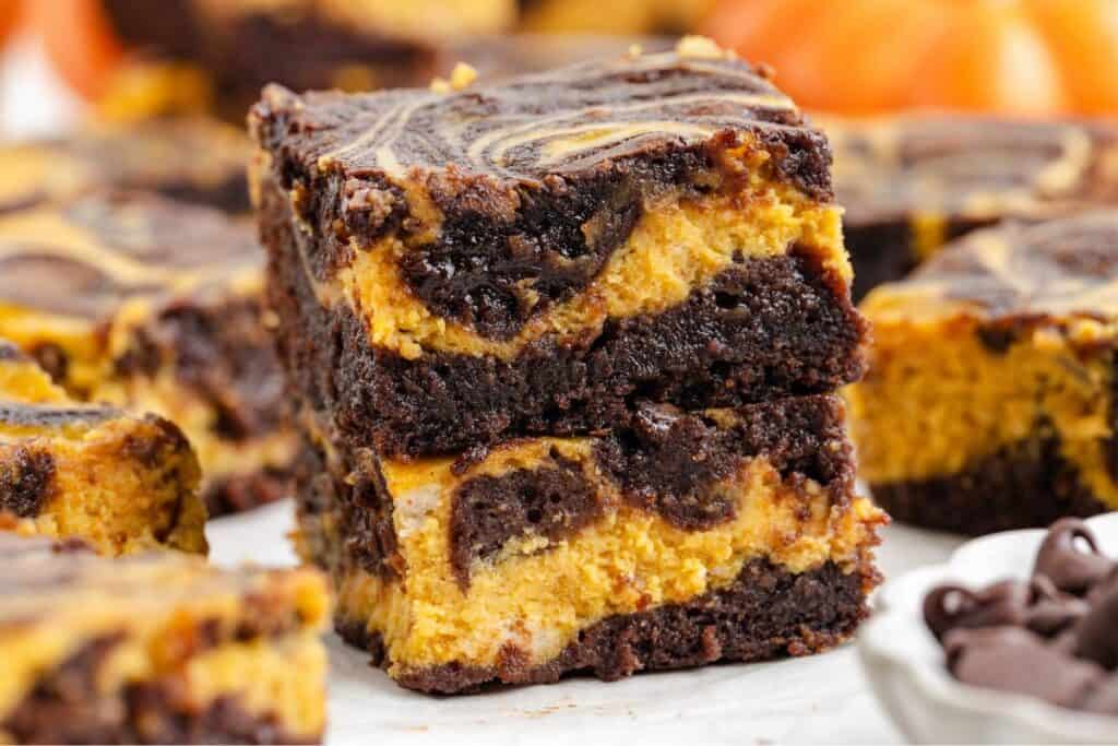 Two stacked pumpkin cheesecake brownies with visible chocolate and orange layers, more brownies in the background.