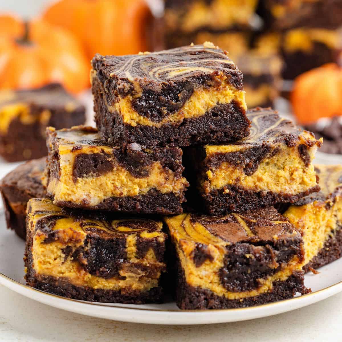 Plate of pumpkin cheesecake brownies, with blurred small pumpkins in the background.
