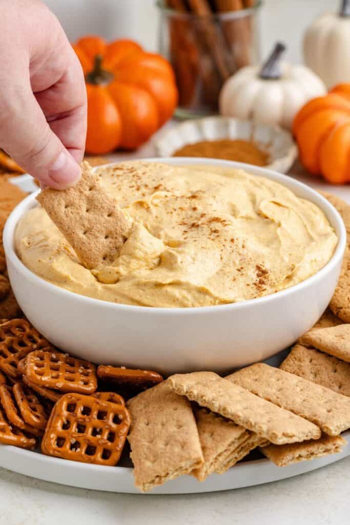 Hand dipping graham cracker into creamy pumpkin pie dip, with more crackers, pretzels, and small pumpkins nearby.