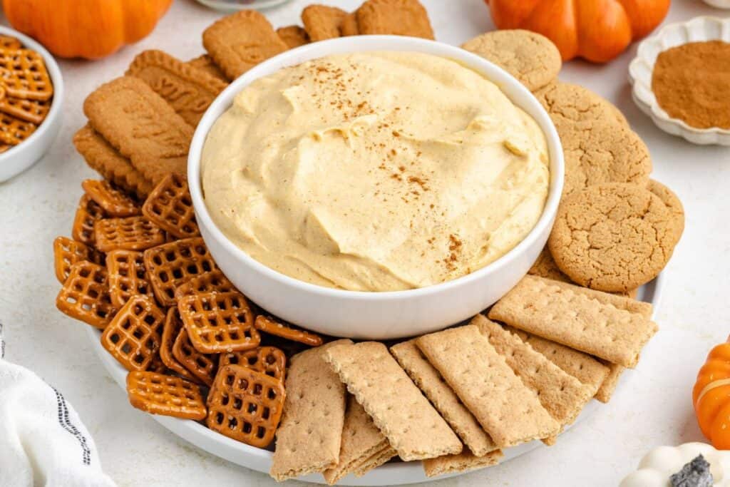 Peanut butter cheesecake dip in a bowl, surrounded by pretzels, graham crackers, cookies, and biscuits on a white plate.