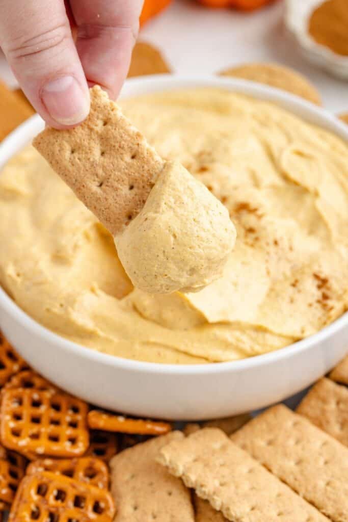 A hand dips a rectangular cracker into pumpkin cream cheese dip, with pretzels and more crackers nearby.