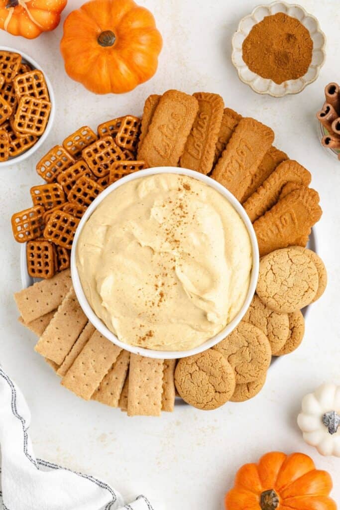 Pumpkin dip in a bowl, surrounded by graham crackers, pretzels, cookies, mini pumpkins, and cinnamon.