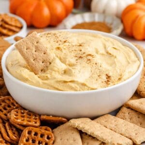 White bowl of pumpkin pie dip with cinnamon, surrounded by graham crackers, pretzels, and small pumpkins in background.