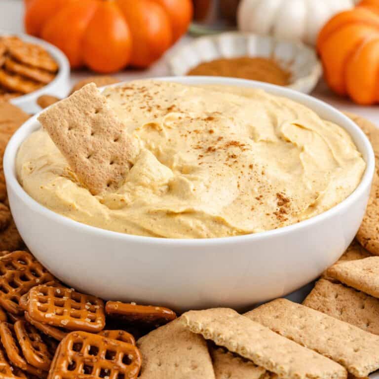 White bowl of pumpkin pie dip with cinnamon, surrounded by graham crackers, pretzels, and small pumpkins in background.