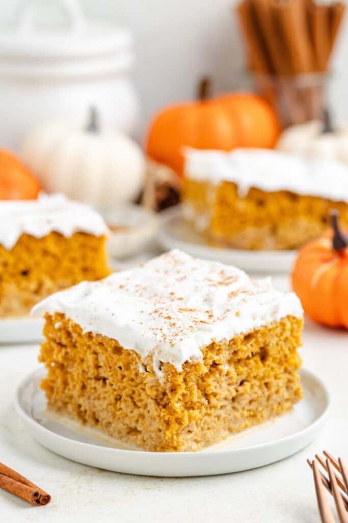 Pumpkin tres leches cake slice with whipped topping on a plate, with extra slices and pumpkins in the background.