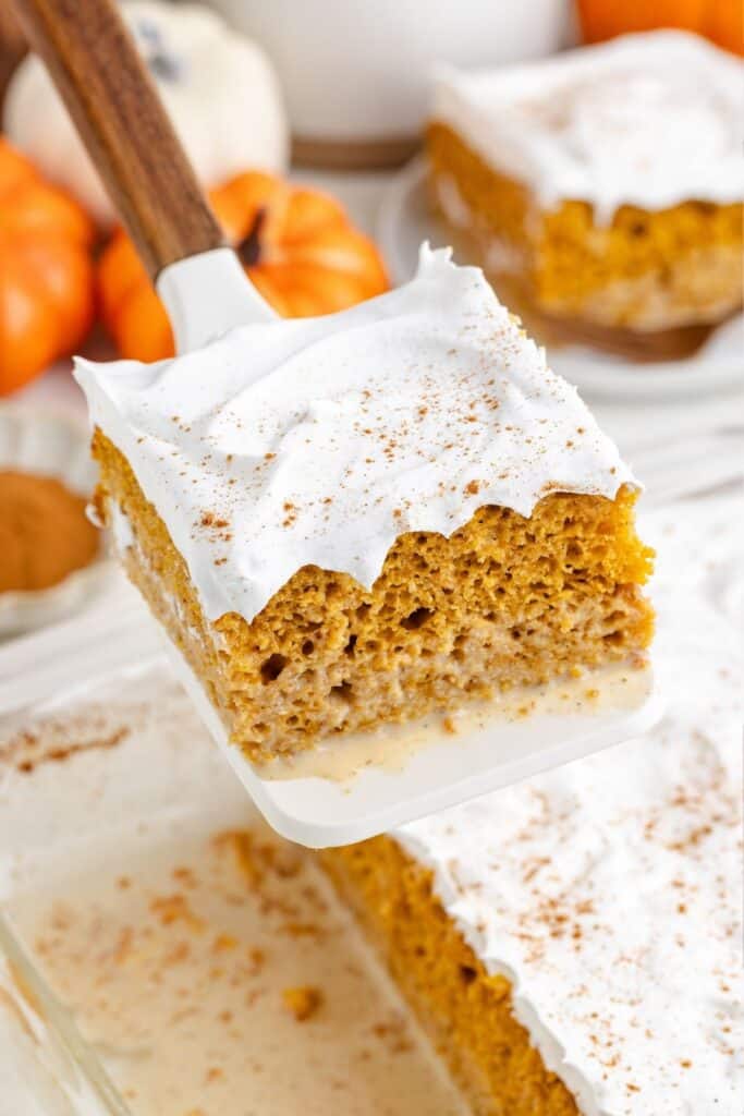 A spatula lifts a pumpkin spice tres leches square with whipped cream; more dessert sits in a glass dish below.