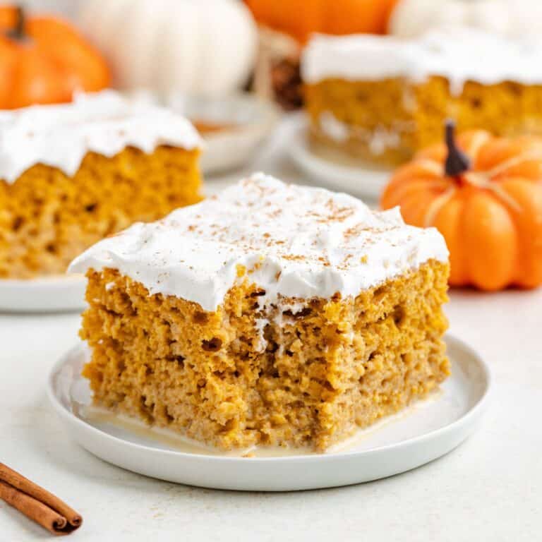 Slice of pumpkin tres leches cake with whipped topping on a plate; pumpkins and another slice in background.