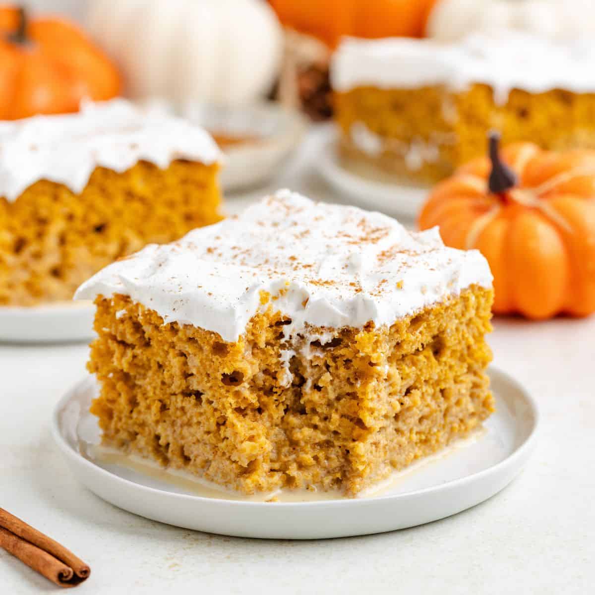 Slice of pumpkin tres leches cake with whipped topping on a plate; pumpkins and another slice in background.