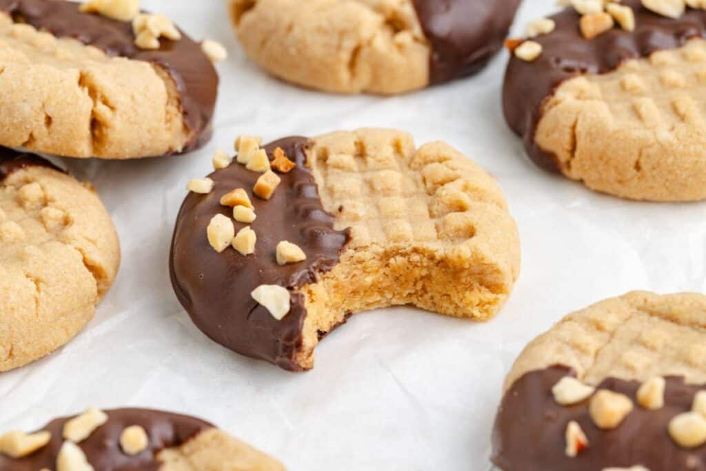 Chocolate-dipped peanut butter cookies with chopped nuts on parchment; one has a bite missing.