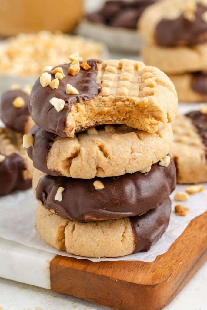 Stack of chocolate dipped peanut butter cookies with chopped peanuts; top cookie has a bite missing.