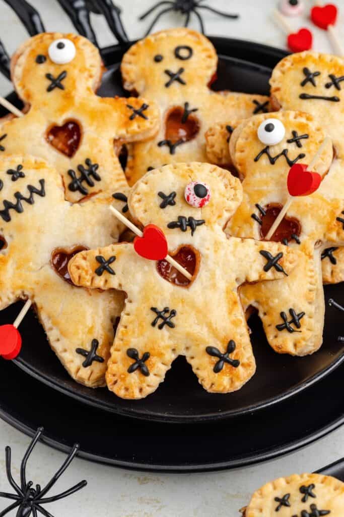 Plate of voodoo doll hand pies with black icing, candy eyes, red heart picks, and nearby plastic spiders.