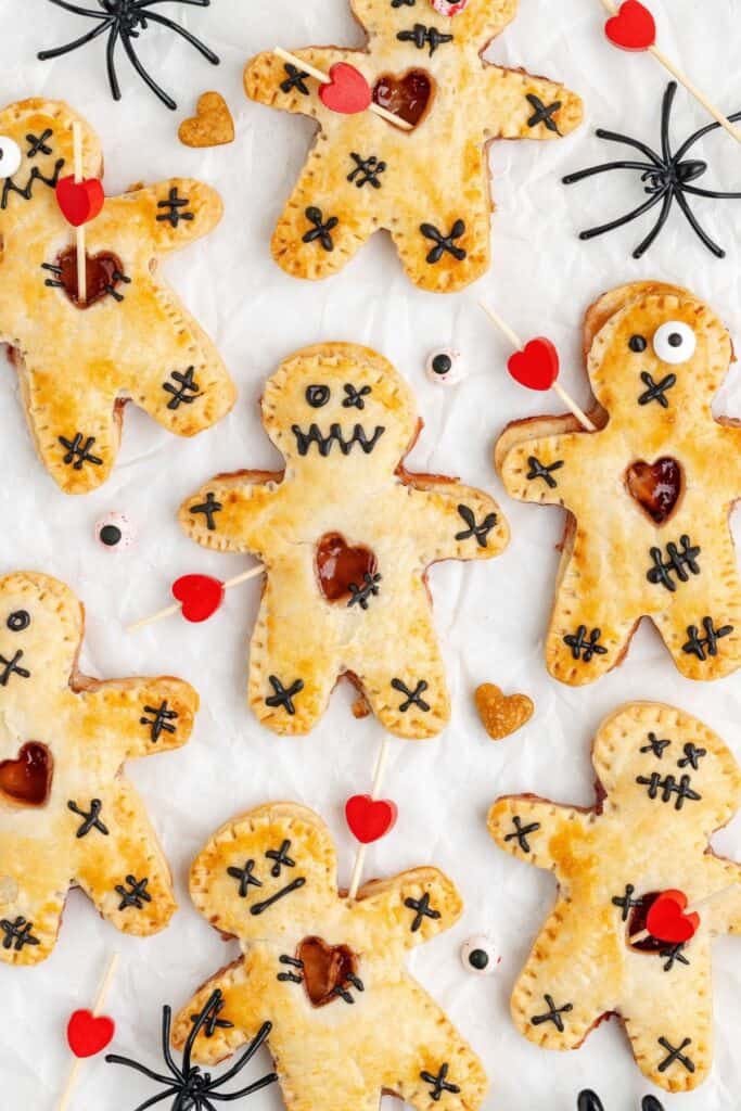 Voodoo doll strawberry hand pies with black icing, heart cutouts, candy eyes, and plastic spiders on white surface.