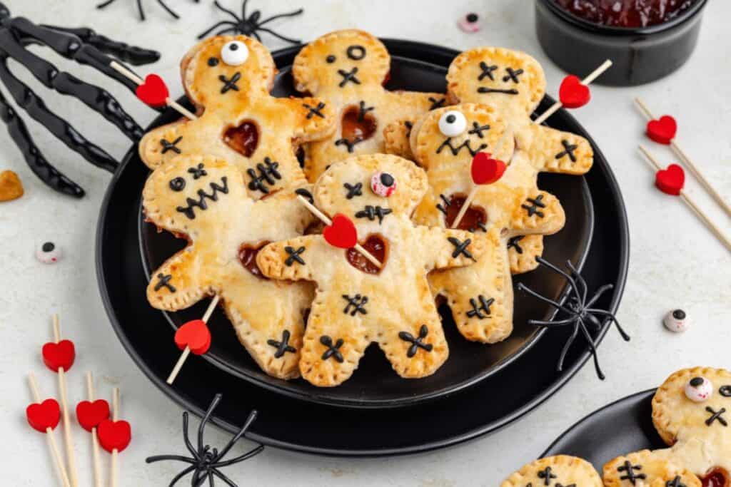 Plate of voodoo doll-shaped pastries with stitched faces, red heart skewers, plastic spiders, and candy eyeballs.