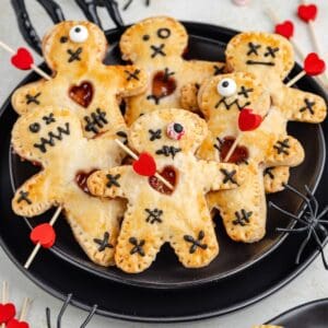 Six voodoo doll hand pies with stitched faces, heart cutouts, and red heart-topped sticks on a plate.