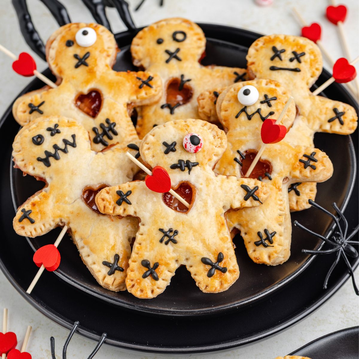 Six voodoo doll hand pies with stitched faces, heart cutouts, and red heart-topped sticks on a plate.