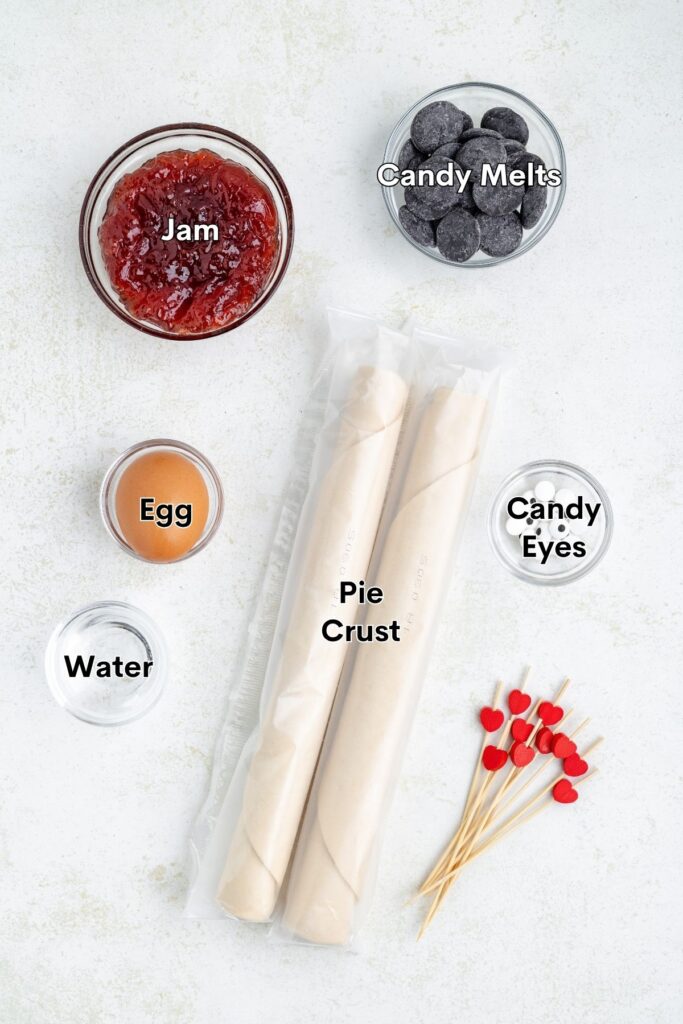 Pie crusts, jam, candy melts, egg, water, candy eyes, and heart sticks on a white surface.