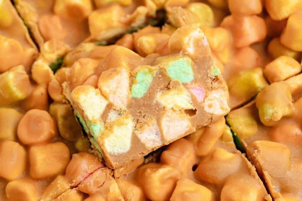 A  peanut butter bar with colorful mini marshmallows, resting atop more marshmallow peanut butter bars.